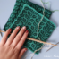 image shows two handknit squares with a leaf motif, in two shades of green. Someone is holding a knitting needle with stitches cast on to it, as if preparing to knit the third square.